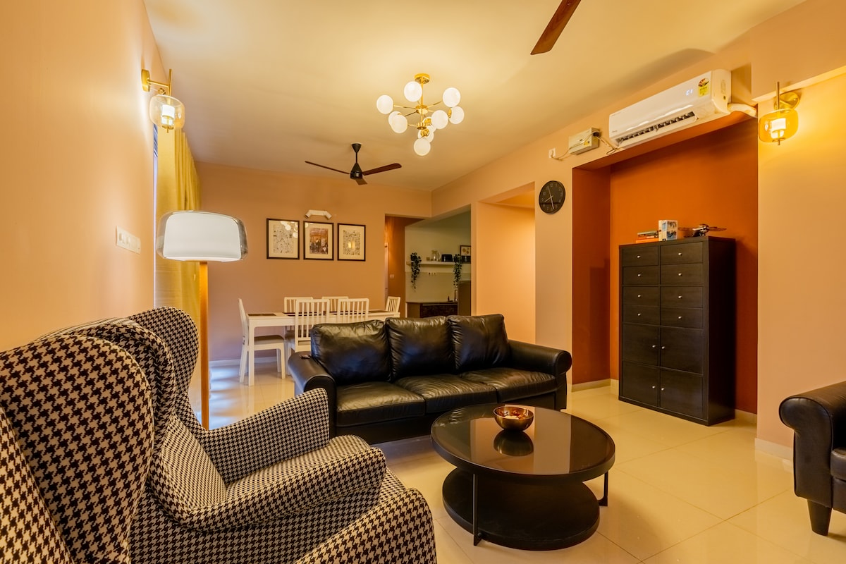 A stylish living area is featured with a mixture of modern and classic furnishings. A checkered armchair sits adjacent to a black leather sofa, complemented by a round coffee table. Warm lighting is provided by a decorative ceiling fixture, while a dining area is subtly visible in the background.