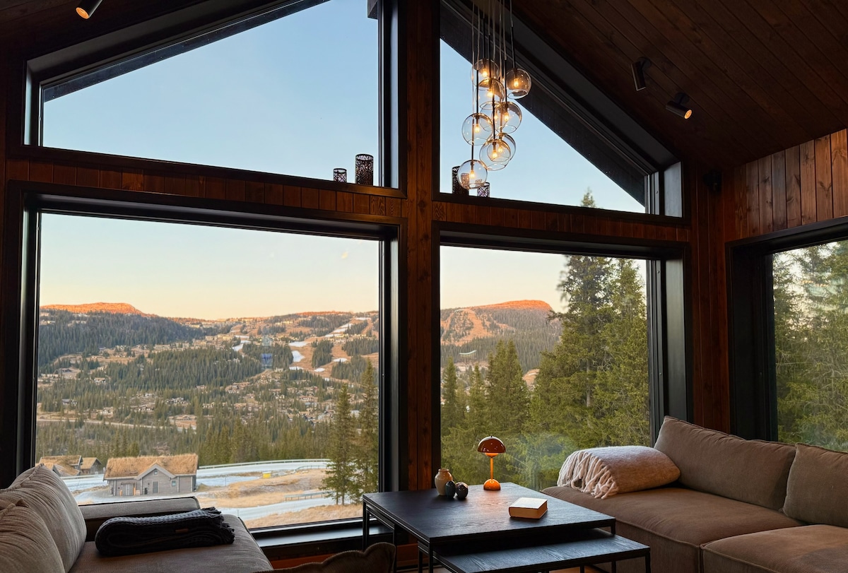 An inviting living area features large panoramic windows that frame expansive mountain views. Plush sofas provide comfortable seating, while a modern light fixture hangs above a simple coffee table. Warm natural wood accents create a cozy atmosphere, enhanced by soft daylight filtering through the glass.