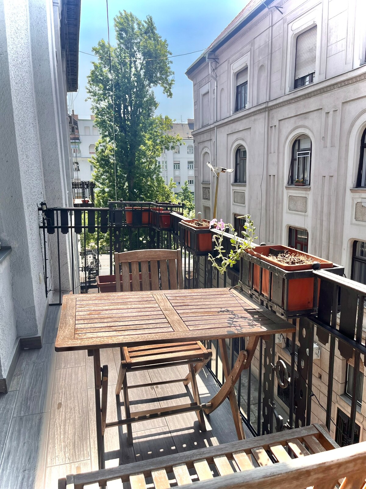 A small balcony features a wooden table and chair set, framed by blooming plants in terracotta pots. Sunlight filters through the surrounding buildings, creating a bright and inviting space for relaxation.