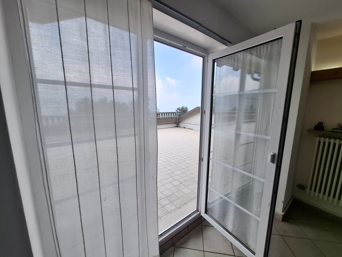 A set of large glass doors opens onto a spacious outdoor terrace, allowing natural light to illuminate the area. Sheer white curtains gently frame the entrance, and the tiled floor extends out, merging the indoor and outdoor environments.
