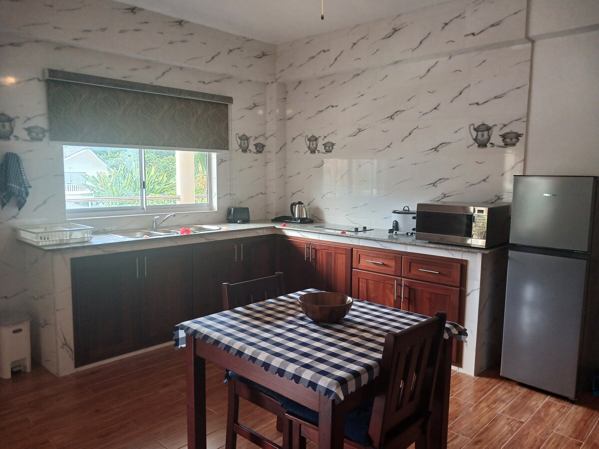 A bright kitchen is equipped with modern appliances including a black microwave and a silver refrigerator. A wooden table is set with a checkered tablecloth and a bowl at the center. Cabinets in a rich wood tone complement the marble-patterned walls and natural light through the window.