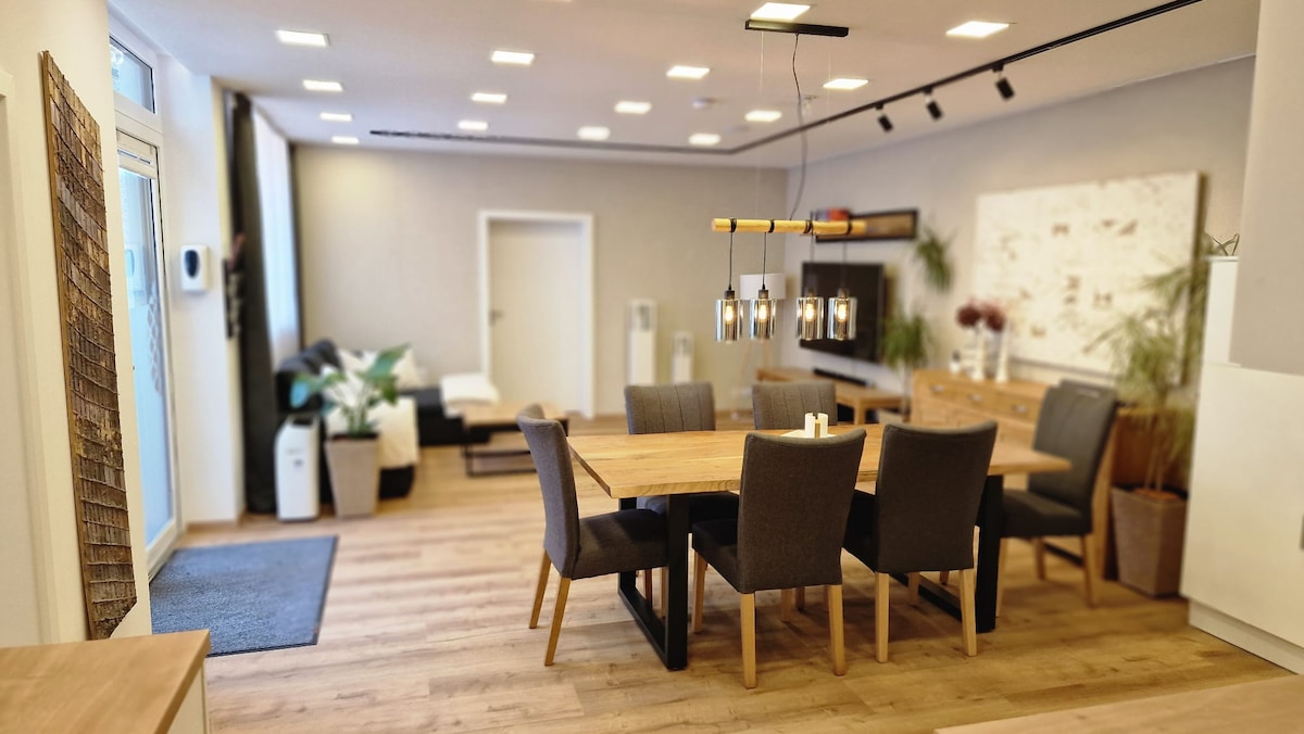 An open living space features a large wooden dining table surrounded by gray upholstered chairs. A comfortable seating area with a dark sofa can be seen in the background. Soft lighting illuminates the room, highlighting decorative plants and modern furnishings.