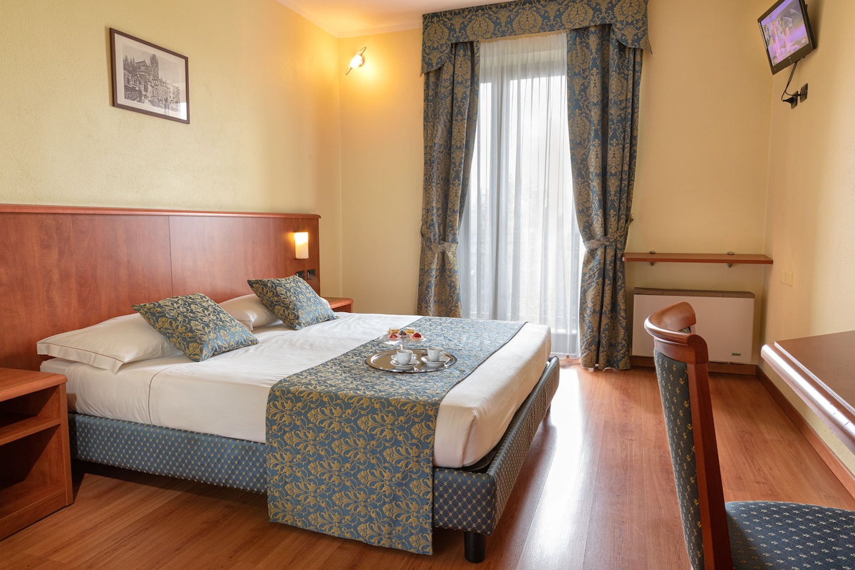 A classic-style bedroom features a spacious bed with decorative pillows. Natural light filters through sheer curtains, illuminating a wooden desk and chair. A framed artwork adds a touch of elegance, while a subtle floral pattern on the bedding complements the room's warm tones.