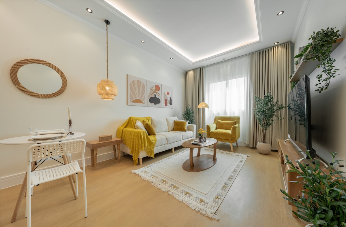 A warm living area features a light sofa paired with a yellow armchair, complemented by decorative wall art. A round coffee table sits on a textured rug, while a small dining table is positioned nearby. Soft lighting enhances the overall inviting ambiance.