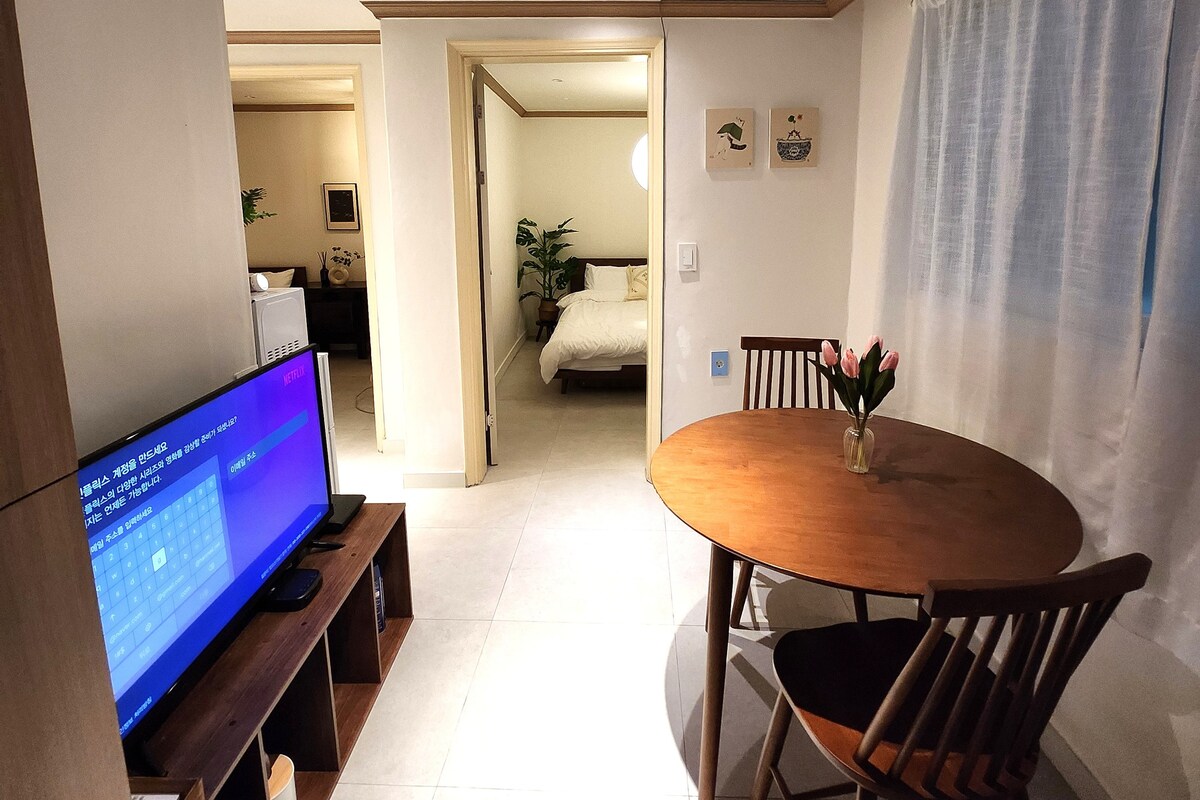 A living area features a wooden dining table accompanied by two chairs. A television is situated on the nearby unit, displaying content. Through the doorway, a glimpse of a bedroom with a bed can be seen, adding to the space's cozy ambiance.