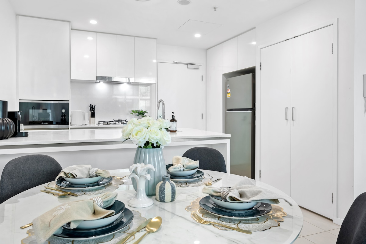 A modern kitchen is featured with sleek white cabinetry and a full suite of appliances, including a microwave and refrigerator. A round dining table is set for four with elegant dishware, showcasing a centerpiece of white flowers. Soft lighting enhances the inviting atmosphere.