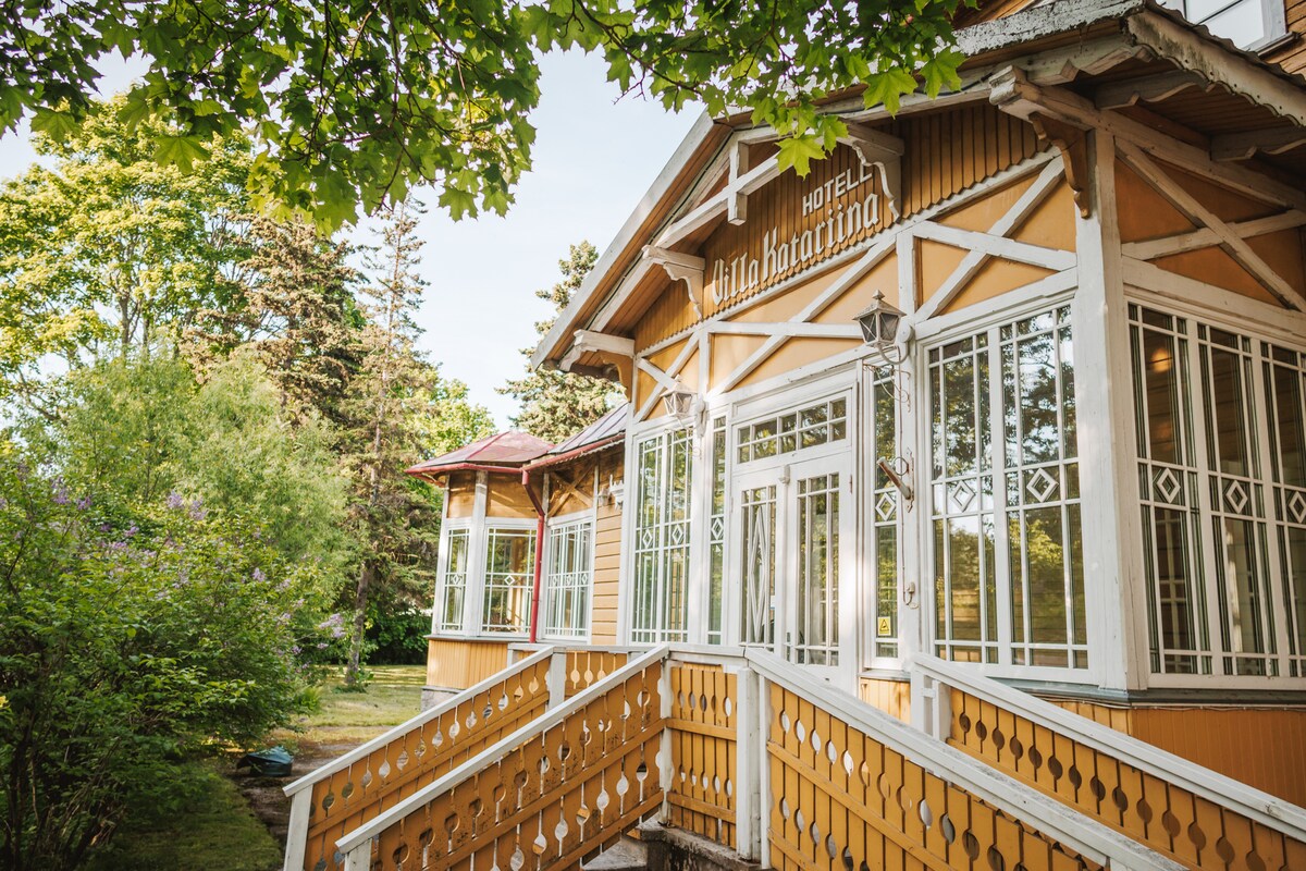 A charming building with wooden architecture features large glass windows framed by white accents, allowing natural light to fill the interior. Surrounded by lush greenery, the entrance is marked by a series of welcoming steps leading into a spacious porch area.