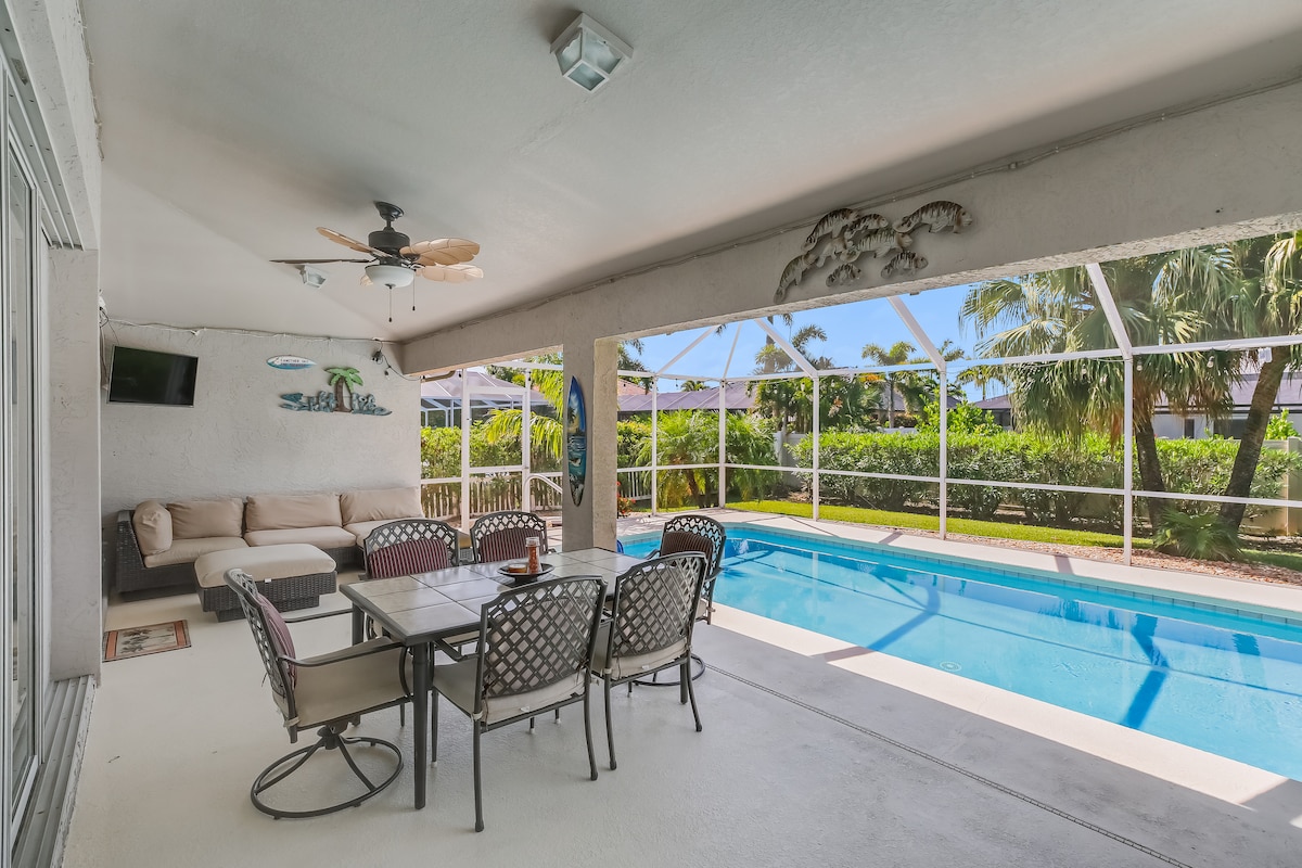 A screened lanai features comfortable seating and a dining table, providing an inviting space for relaxation. A private pool extends into a lush outdoor area, with palm trees visible beyond the pool enclosure, offering a serene atmosphere.