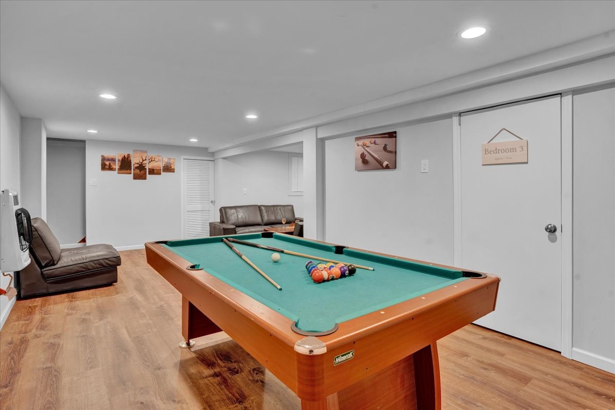 A game room is designed with a pool table as the focal point, accompanied by a couch positioned along one wall. Artwork is displayed on another wall, while two doors lead to adjoining spaces. The room features wooden flooring and recessed lighting.