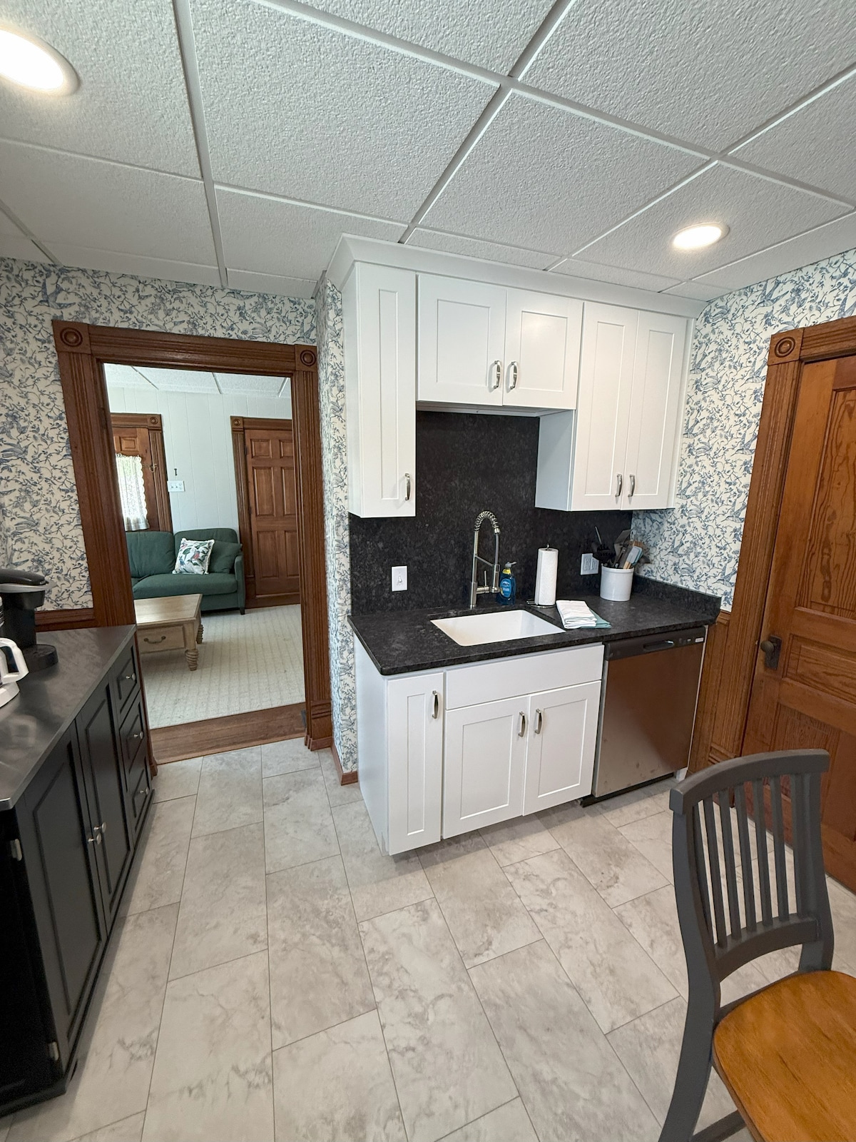 A functional kitchen area is presented with modern white cabinetry and a dark stone countertop. A stainless-steel sink and dishwasher are visible, alongside essential kitchen appliances. Light-colored tile flooring complements the walls adorned with floral wallpaper, creating a clean and organized space.