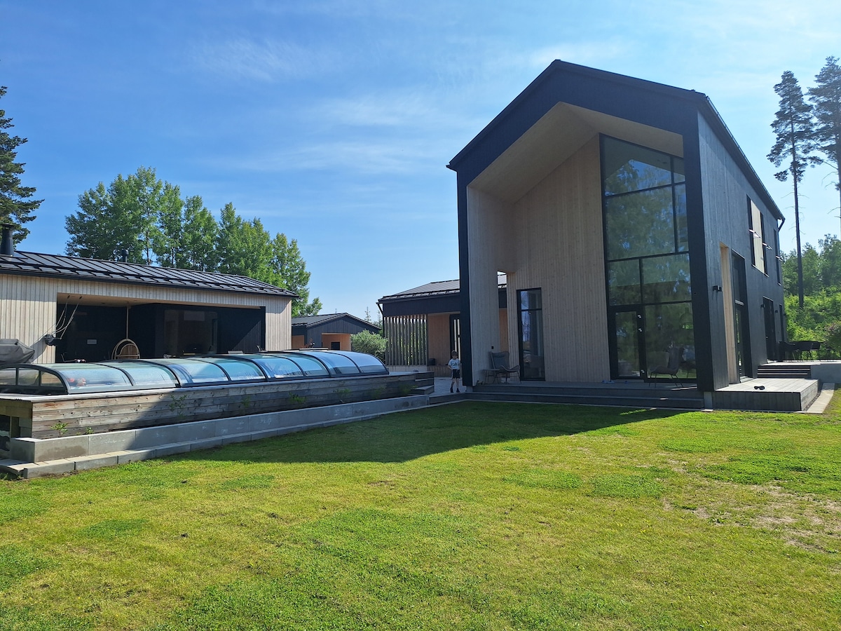 The exterior of the villa is showcased, highlighting a modern structure with large glass windows. A covered swimming pool is situated in the foreground, surrounded by well-maintained grass. Trees line the background, complementing the tranquil environment.