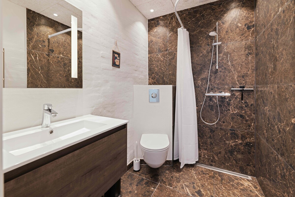 The bathroom features dark marble walls and a sleek, modern design. A vanity with a white sink and a minimalistic faucet is installed. A shower area with a handheld showerhead is enclosed by a white curtain. A wall-mounted toilet is visible, contributing to the contemporary aesthetic.