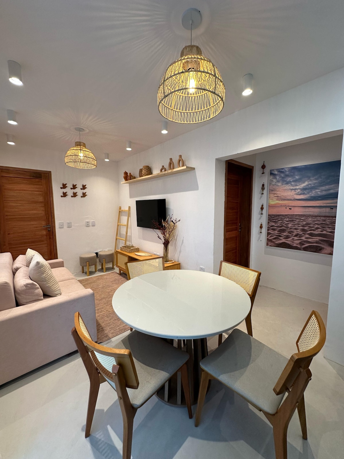 A modern living space is highlighted by a circular dining table with four light-colored chairs. Soft lighting from woven pendant lamps creates a gentle ambiance. A comfortable sofa sits against the wall, and a decorative shelf above a television displays a few ornamental pieces.