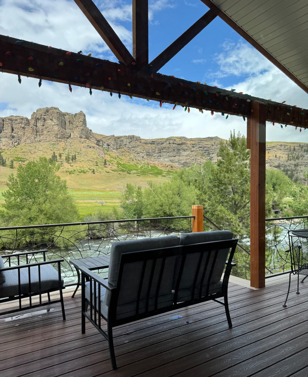 A covered patio is shown with a comfortable seating area overlooking the Stillwater River. Lush greenery and rocky cliffs are visible in the background, while decorative string lights are draped along the wooden beams above, adding a welcoming touch.