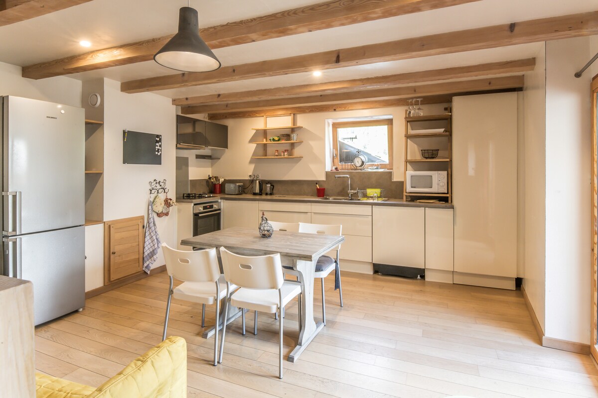 The kitchen space features modern appliances, including a stainless steel refrigerator and an oven. A central dining table is surrounded by four white chairs, while open shelving displays kitchenware. Wooden beams add warmth to the overall design, complemented by natural light entering through the window.