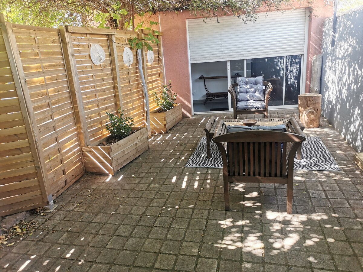 A private outdoor space is showcased with a paved patio area. Wooden planters with greenery border the space, and a cozy seating arrangement is positioned on a decorative rug. Natural light filters through the overhead canopy, creating a serene atmosphere.