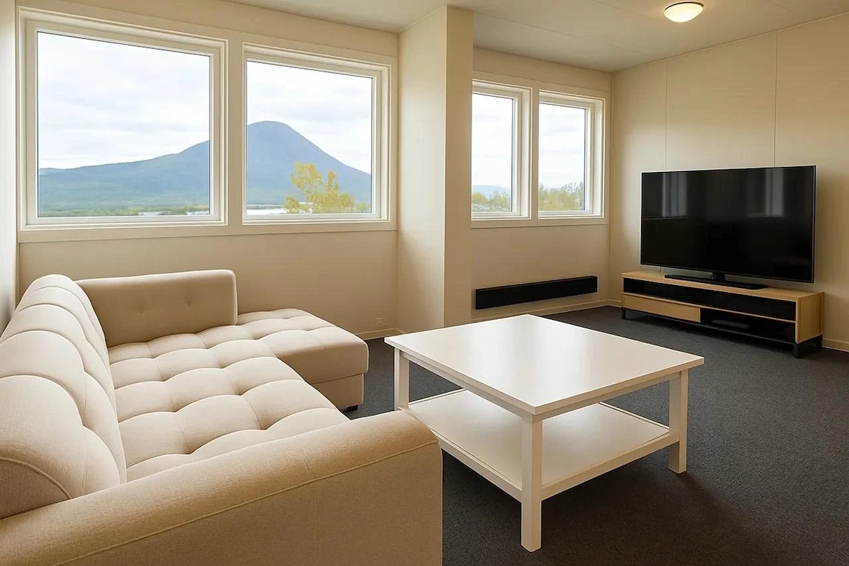 A light-filled living area features a spacious beige sectional sofa positioned near large windows, offering views of a distant mountain. A simple white coffee table is centered in front of the sofa, while a black media console and television are located against the opposite wall.