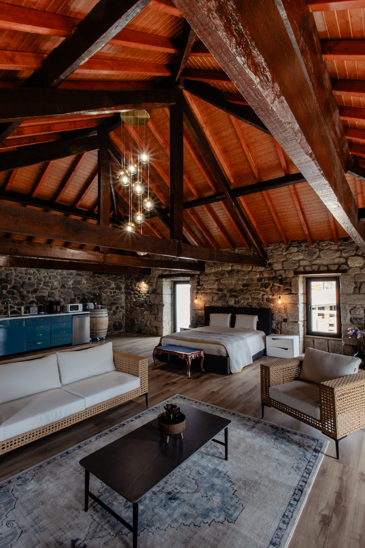 A spacious interior features exposed wooden beams and a rustic stone wall. A comfortable seating area is arranged with light-colored sofas and a dark table. A neatly made bed is set against a window, allowing natural light to illuminate the room.
