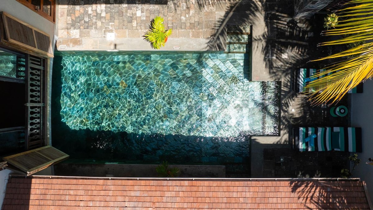 An aerial view showcases the private infinity pool with clear, shimmering water reflecting sunlight. Surrounding the pool are stone pathways and lounge chairs, framed by tropical palm leaves, creating a serene and inviting outdoor space.