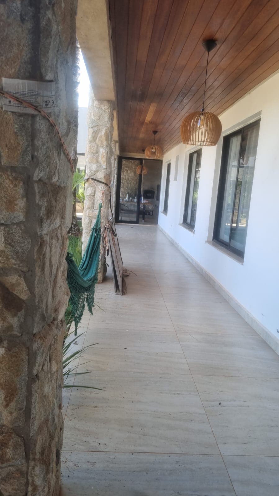 A covered porch is visible, featuring stone pillars and wooden ceiling accents. Hanging light fixtures add a warm touch. Expansive windows allow natural light, and a hammock is positioned along the side, contributing to a relaxed atmosphere. The flooring is made of light-colored tiles.