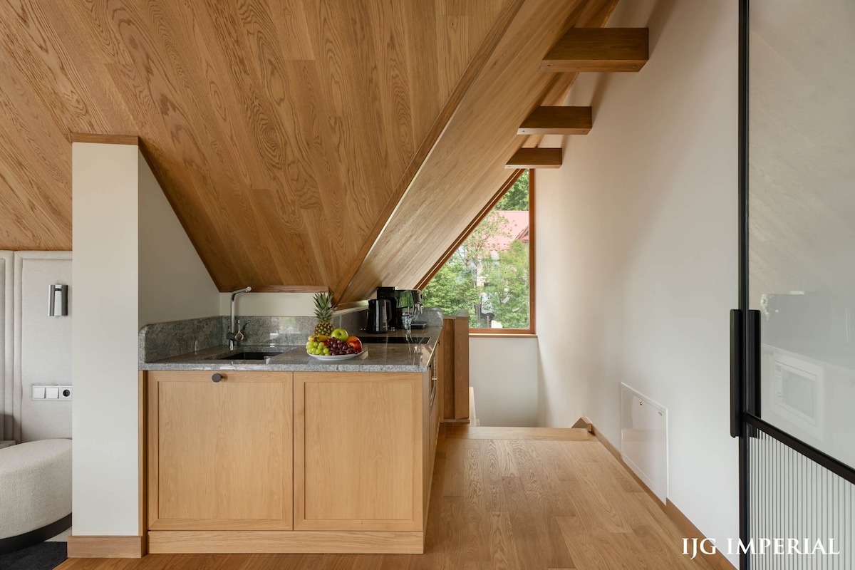 A modern kitchenette features wooden cabinetry and a granite countertop. A fruit bowl filled with various fruits is displayed, alongside a coffee maker and kettle. Natural light filters through a window, enhancing the warm wood tones of the ceiling and floor.