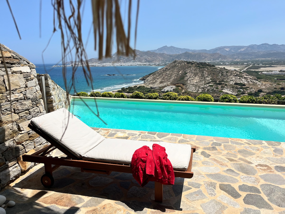 A private pool with a stunning view of the Aegean Sea is framed by natural stone walls. A sun lounger with a light cushion rests nearby, complemented by a neatly folded red towel. Rolling hills and coastal scenery create a tranquil backdrop.