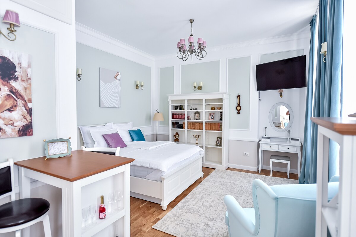 A charming guestroom features a well-made spacious bed against light green walls. A bookshelf filled with books is placed nearby. A television is mounted on the wall, and a cozy seating area is arranged with a light blue chair. Natural light enters through a window dressed in flowing curtains.