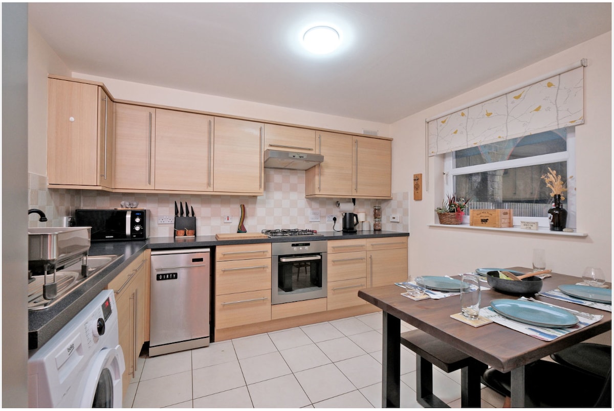 The kitchen features light wood cabinetry, an integrated oven, and stainless steel appliances. A dark rectangular dining table is set with dishes and glassware. A window allows natural light to enter, highlighting the tiled flooring and overall functionality of the space.