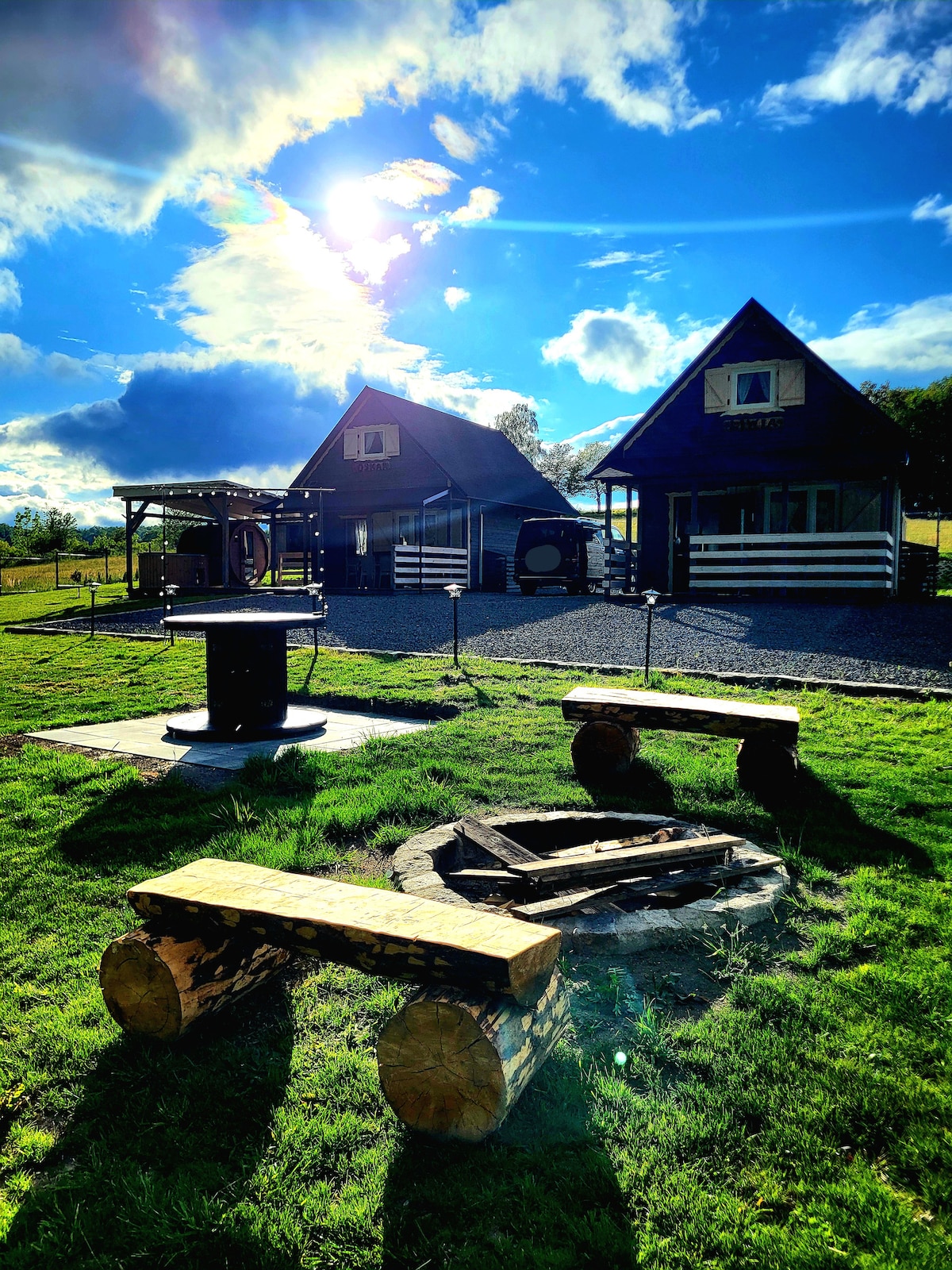 Two charming cabins are positioned in an open grassy area, accompanied by a fire pit constructed from logs. Seating made from similar logs surrounds the fire pit. A clear blue sky with scattered clouds enhances the natural setting, complemented by soft sunlight illuminating the scene.