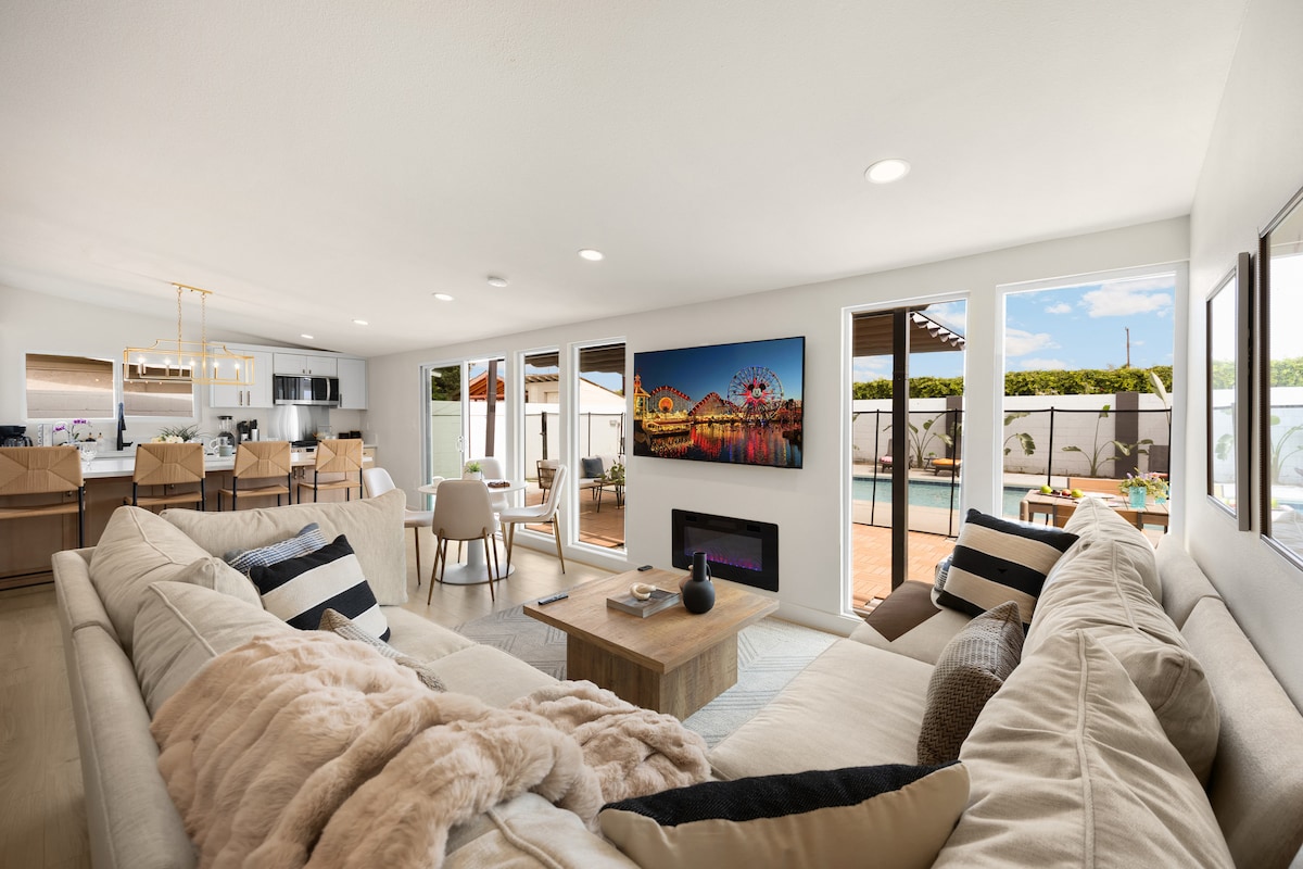 A spacious living area features modern furnishings with a large modular sectional sofa and a sleek electric fireplace. Bright sunlight enters through multiple windows, offering views of outdoor seating and a pool area. A flat-screen TV displaying a colorful theme park scene adds a touch of entertainment.