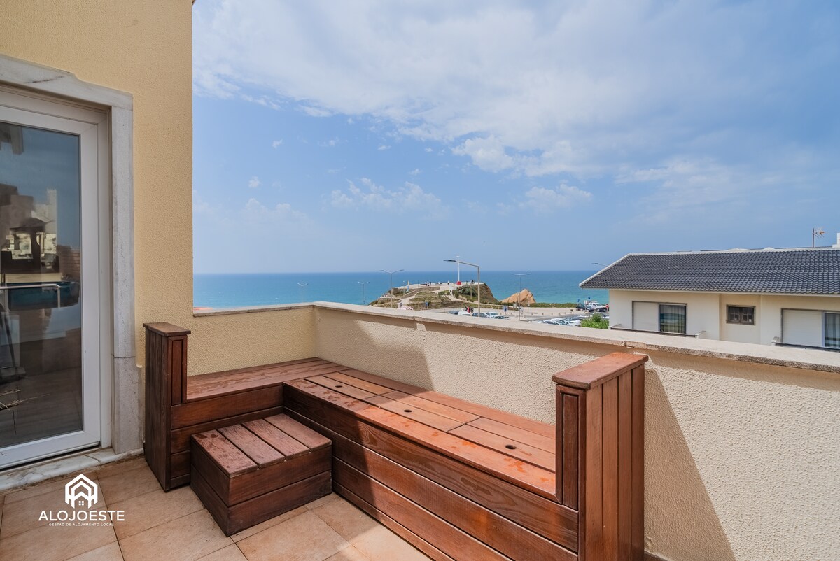 A private terrace features wooden seating that provides a relaxed area to enjoy the ocean views. The expansive sea can be seen in the distance, under a partly cloudy sky, and nearby structures are visible along the shoreline.