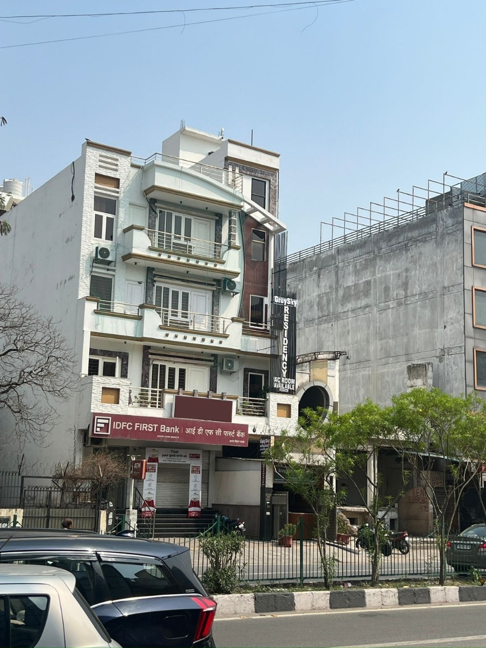 A multi-story building is displayed with a clean, modern façade. The ground floor features a bank entrance, while several air conditioning units are visible on the upper levels. Surrounding greenery enhances the urban landscape, and parked cars line the street in front.