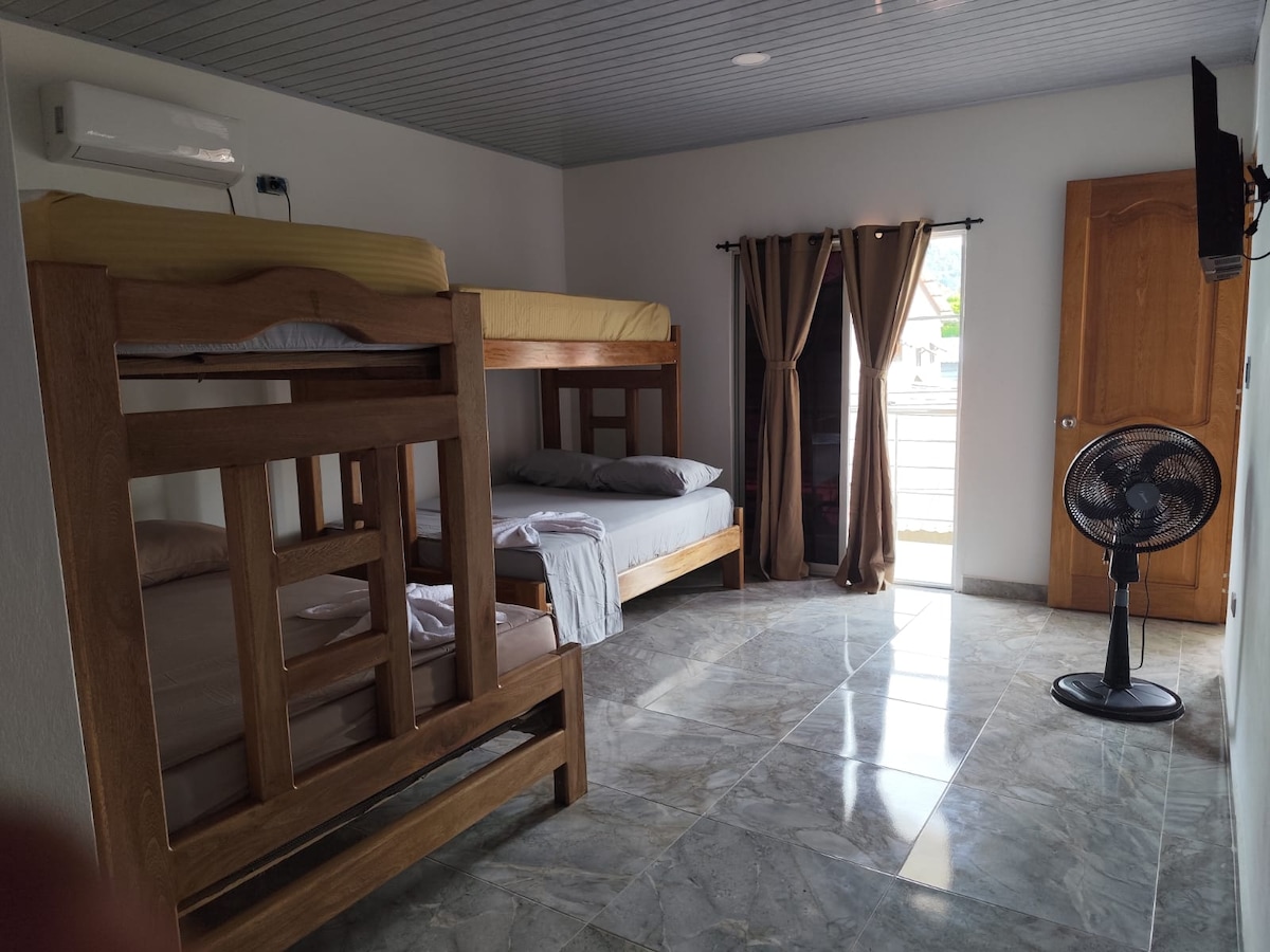 A spacious room features a wooden bunk bed alongside a double bed. Natural light fills the space through an open door, illuminating the polished marble floor. A ceiling fan is positioned near the entrance, and curtains frame the door, adding a touch of warmth to the room.