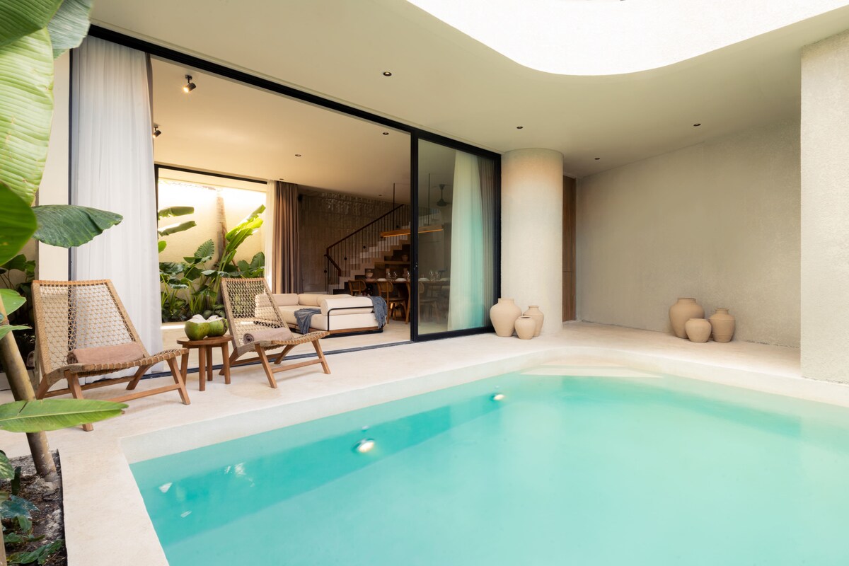 A serene outdoor space features a private pool with clear water, surrounded by smooth concrete. Comfortable seating with natural elements is visible, complemented by potted plants. Large glass doors allow natural light to flow into the indoor area, enhancing the connection between indoors and outdoors.
