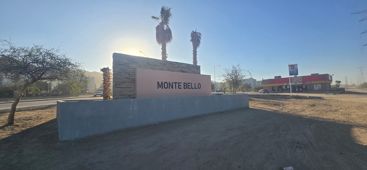 A large sign displaying 'MONTE BELLO' stands prominently, surrounded by palm trees. The area appears sunlit with a clear sky, and a commercial building is visible in the background, providing context to the location.