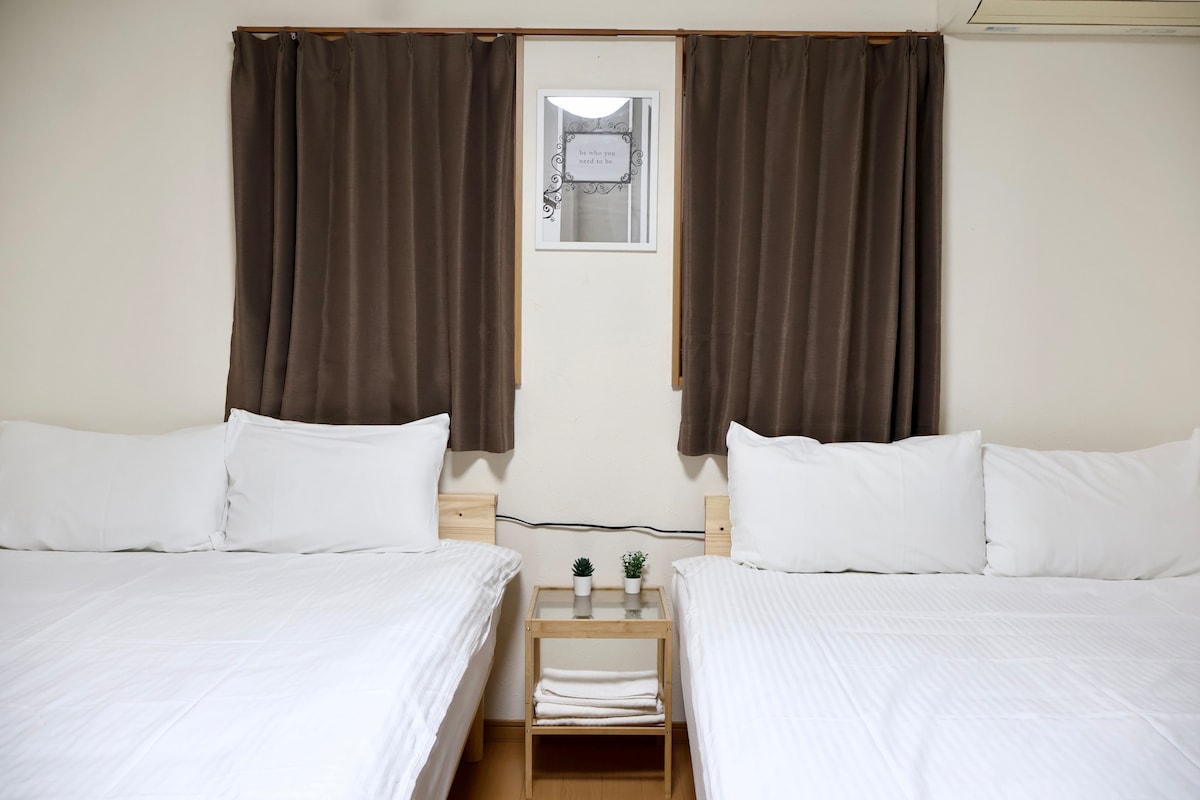 Two spacious beds are placed side by side, each adorned with white linens and multiple pillows. A small wooden nightstand is positioned between them, featuring decorative plants and towels. Natural light filters through the framed window above, covered by brown curtains.