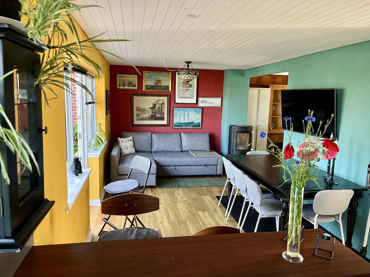 An inviting living area features a gray sofa against a red accent wall, complemented by various framed artworks. A dining table surrounded by light chairs is visible, with a tall vase of flowers adding a touch of greenery. Natural light enters through the window.
