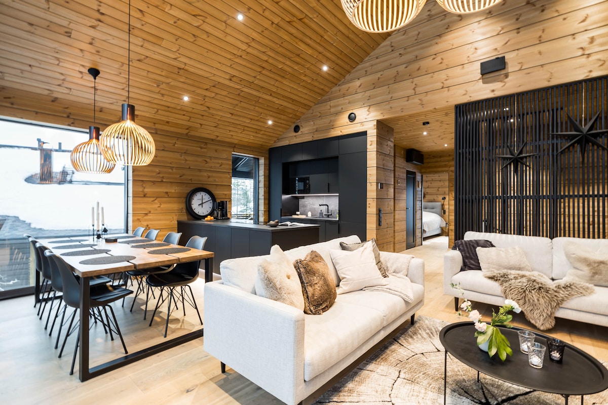 A modern open-plan living area features high wooden ceilings and large windows showcasing a scenic view. A spacious sofa is positioned near a black coffee table, while a dining table with stylish chairs is set for meals. Unique pendant lights add a contemporary touch.