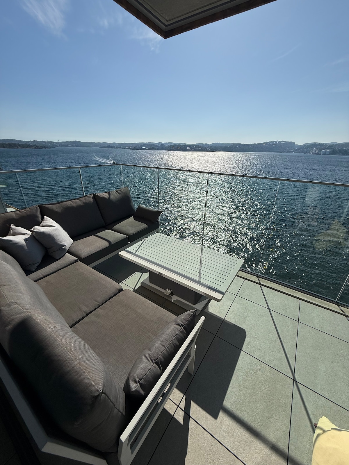 A spacious balcony is furnished with a comfortable outdoor sofa and a glass-top coffee table, offering views of the shimmering water. The clear blue sky reflects on the surface of the sea, providing a calming backdrop. Sunlight brightens the area, creating an inviting atmosphere.