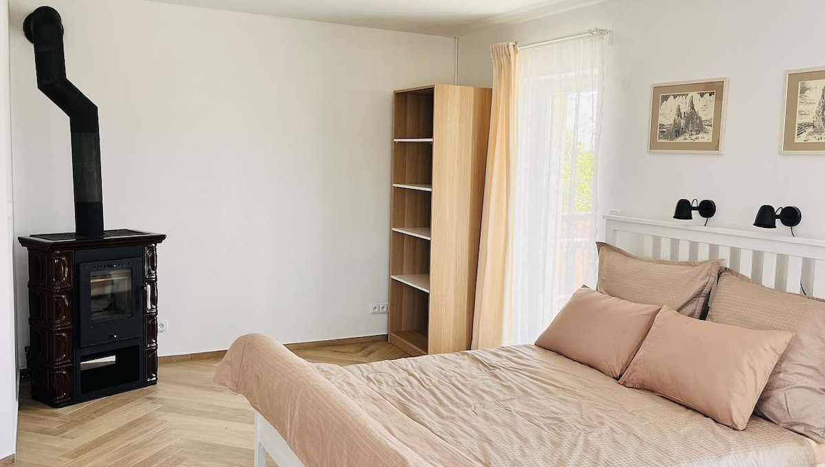 A spacious bedroom features a large bed with soft bedding and two pillows. A wood-burning stove stands against the wall, complemented by a wooden shelving unit and soft curtains that allow natural light to fill the room.