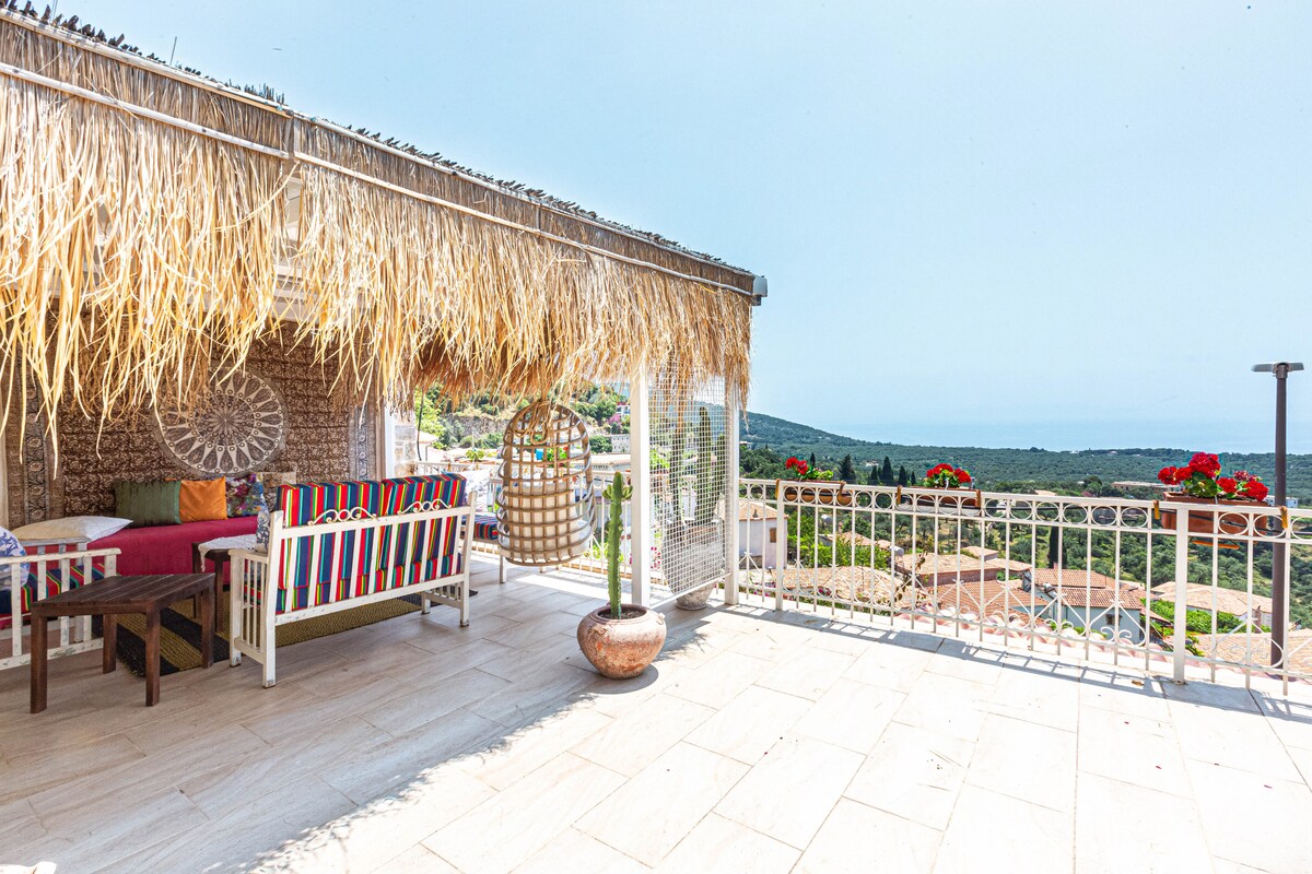 A spacious terrace features a thatched roof, providing shade over colorful seating areas. Planters with greenery are positioned throughout. Panoramic views of the Ionian Sea and surrounding landscape are visible, enhancing the outdoor experience.