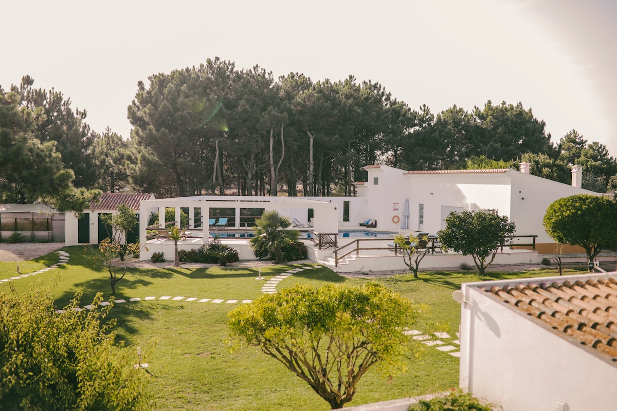 A spacious garden is surrounded by a modern white structure, featuring a pool and relaxing outdoor areas. Lush greenery provides a serene backdrop, while pathways lead through well-maintained lawns. The setting emphasizes tranquility, with trees visible in the distance.