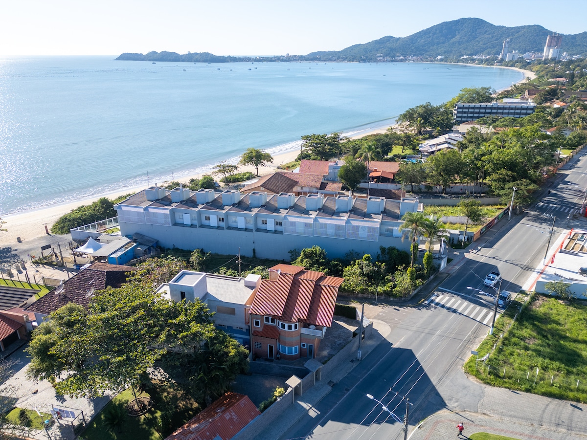 An aerial view captures the proximity of the property to the beach, with clear blue waters stretching along the coastline. The surrounding area features a mix of greenery and buildings, highlighting the accessibility to local amenities and the serene environment near the shore.
