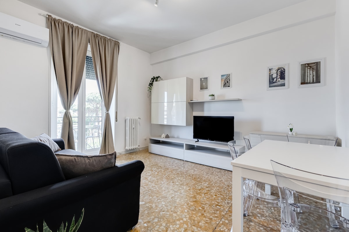 A bright living area features large windows with light curtains, allowing natural light to fill the space. A modern television unit is positioned against a white wall, while a sleek dining table with transparent chairs complements the contemporary design. Comfortable seating is provided by a dark couch.