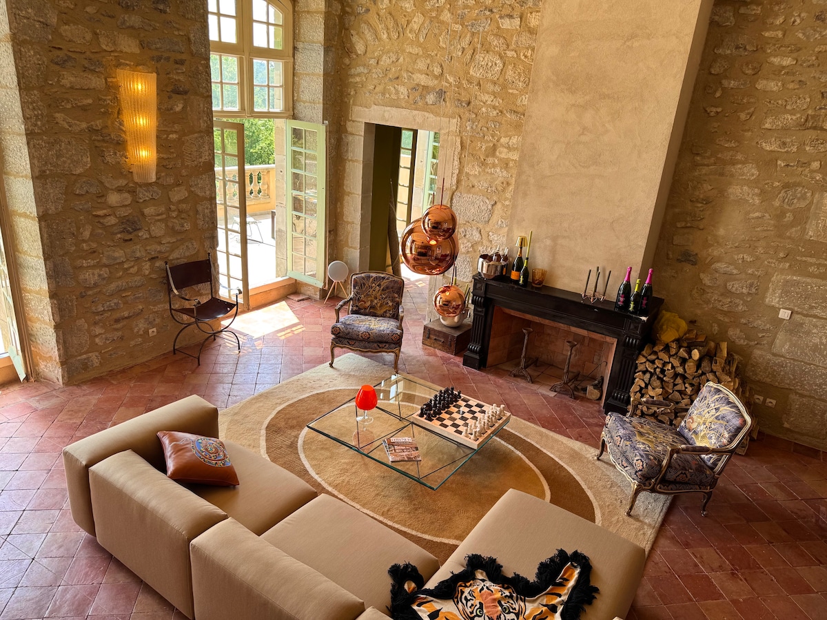 A spacious living area features stone walls and high ceilings, complemented by large windows allowing natural light. A comfortable sectional sofa sits on a circular rug, while a glass coffee table displays a red balloon and a chess set. Stylish armchairs are positioned nearby.