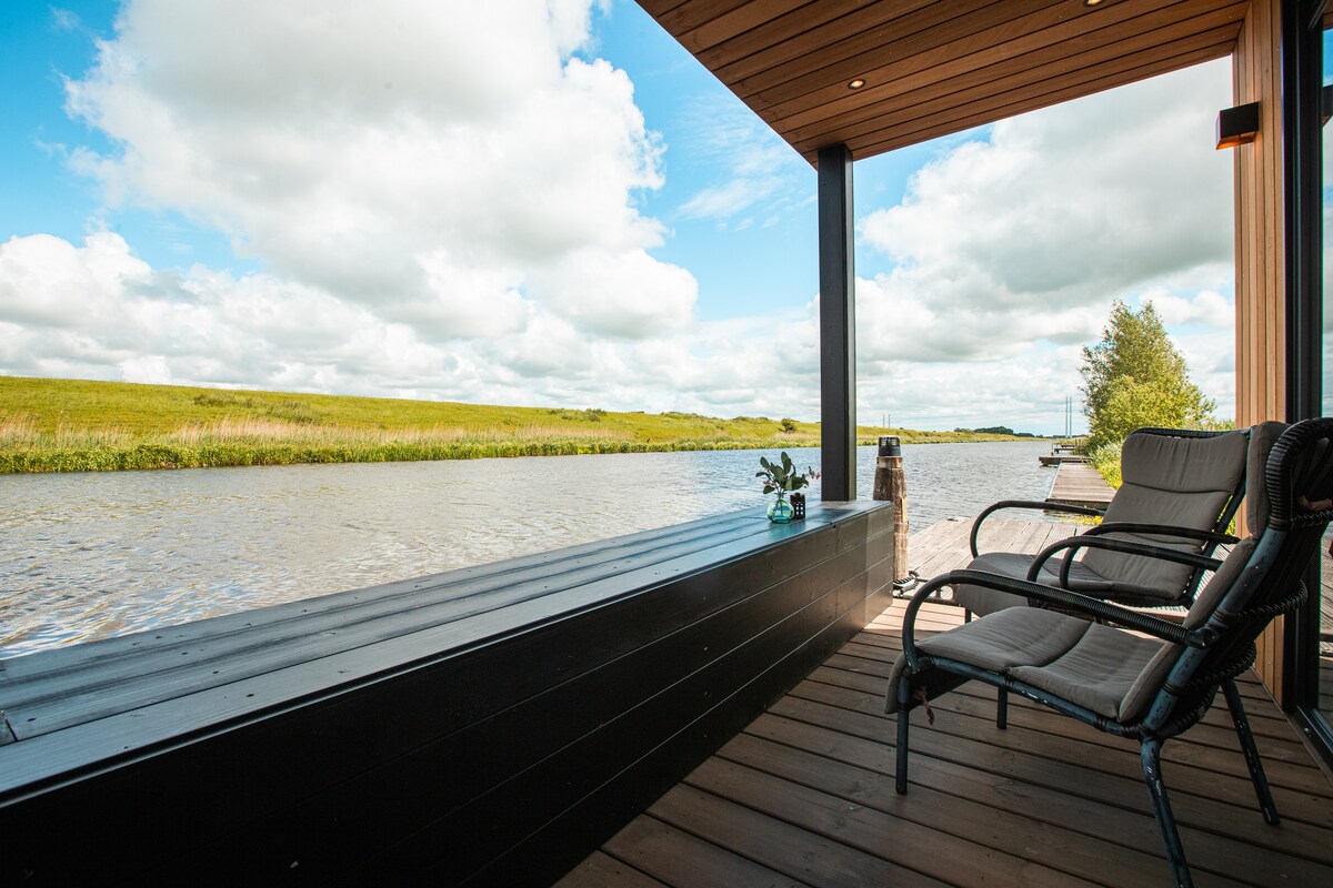 A serene outdoor seating area features two chairs positioned on a wooden deck overlooking the calm water. Lush green fields stretch into the distance under a partly cloudy sky, creating a peaceful atmosphere for relaxation.