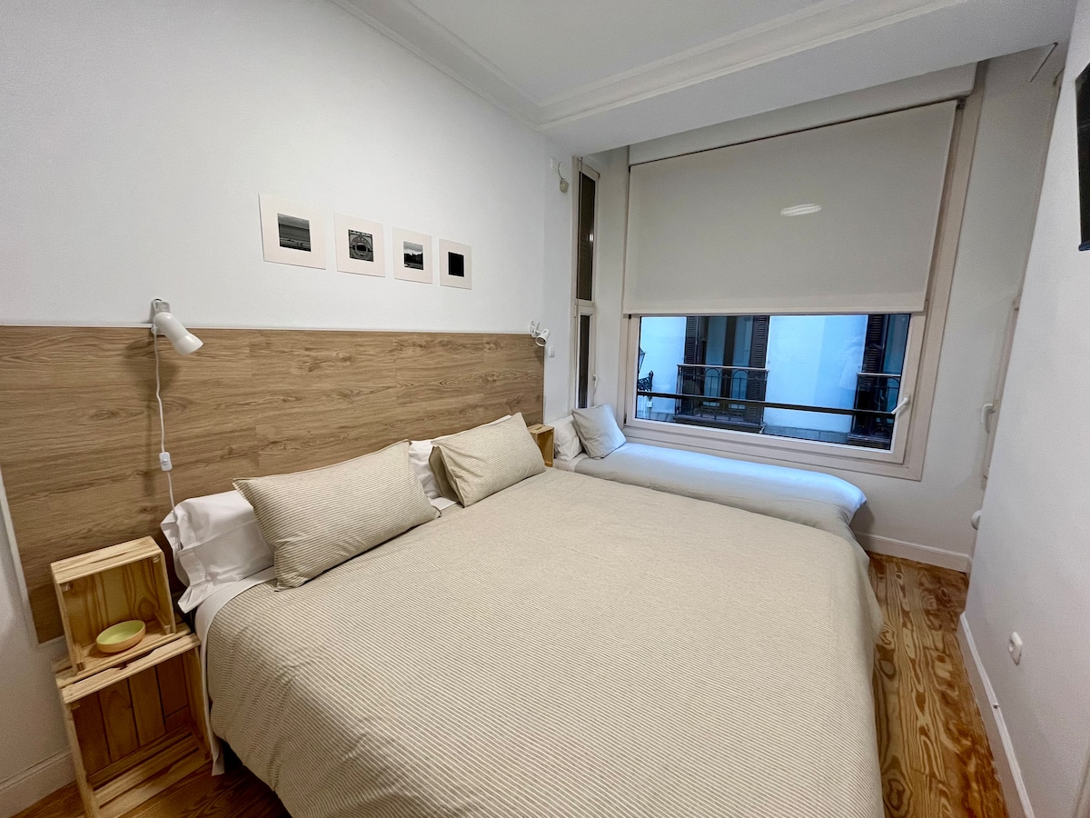 The sleeping area features two spacious beds, each adorned with neutral-colored linens and ample pillows. A light wood headboard complements the wooden floor, while framed artwork hangs above the beds. Natural light fills the room through a large window with blinds.