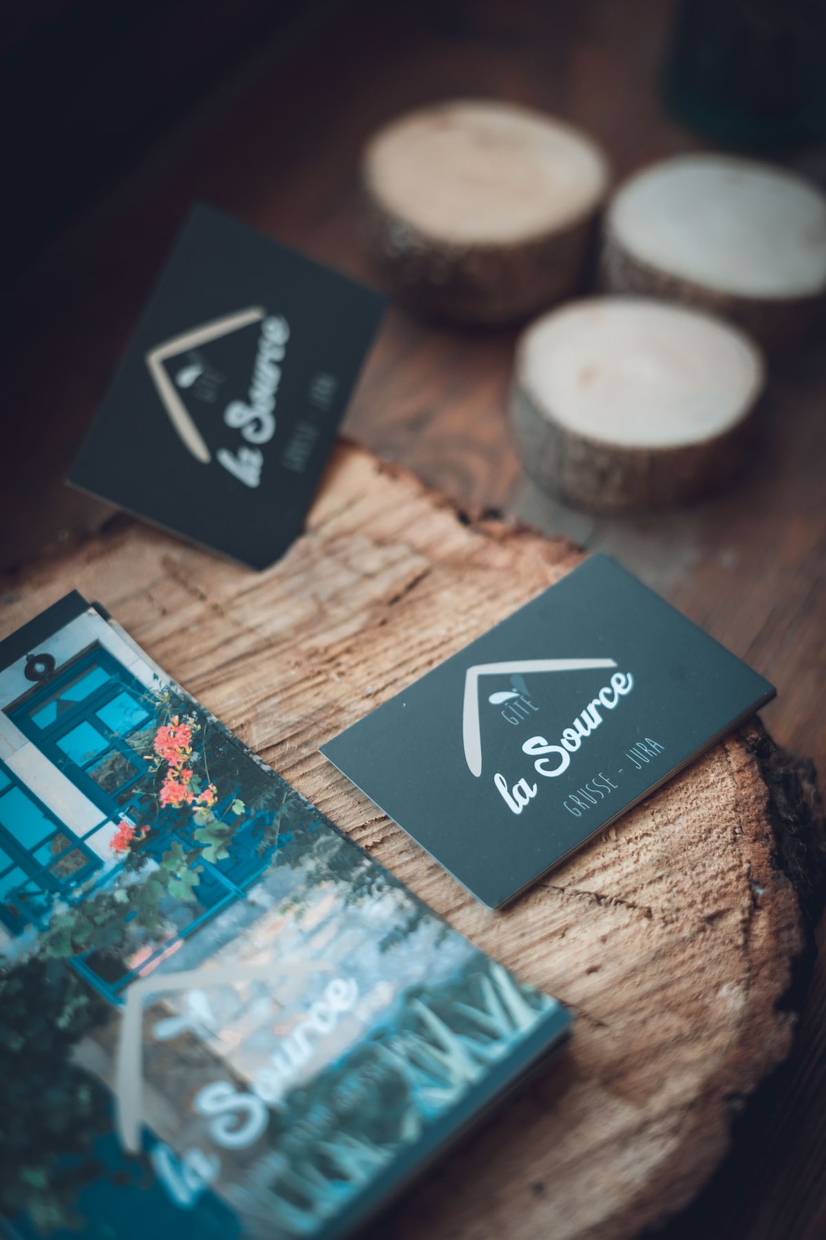 Business cards for 'gîte la Source' are displayed atop a rustic wooden surface, accompanied by circular wooden coasters. A booklet featuring vibrant imagery and information about the property can be seen nearby, enhancing the welcoming ambiance of the space.
