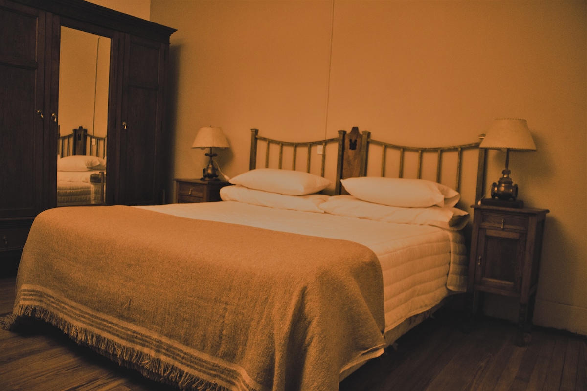 A warm-toned bedroom features a spacious bed topped with neatly arranged pillows and a textured blanket. Wooden nightstands with lamps flank the bed, and a large mirror reflects the cozy decor. A wooden wardrobe adds storage space, enhancing the room's functionality.