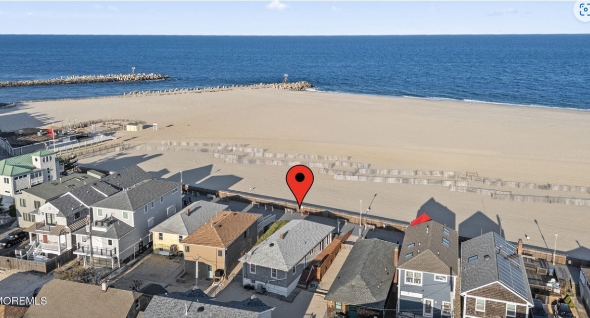 An aerial view captures the home's location along Point Pleasant Beach, with the vast Atlantic Ocean stretching in the background. The sandy beach is visible, complemented by a jetty in the distance. Nearby buildings line the boardwalk, showcasing a coastal community.